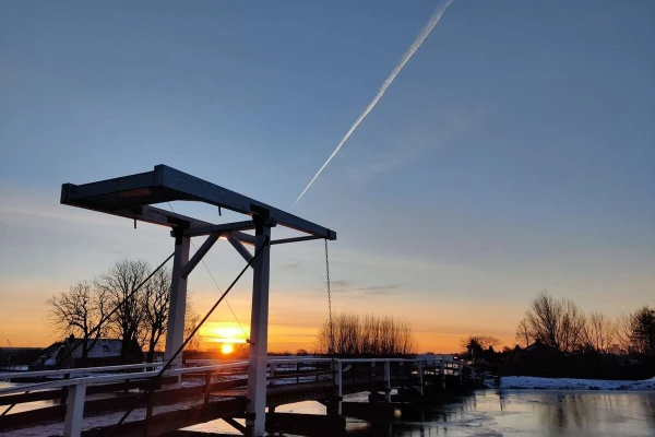 Sunrise through a classic bridge over a frozen canal. In the blue sky a white stripe is seen from a plane.
